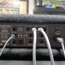 Zadní strana kytarového komba v černém textilním potahu. Na šedém kovovém panelu je nápis CARVIN R600 a dále označení FS-22 FOOTSWITCH, GRAPHIC EQ, E…