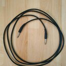Černý instrumentální kabel s úhlovým a rovným jack konektorem. Kabel je svinutý do kruhu na světlém dřevěném povrchu. Na kabelu ani konektorech nejso…