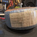 Fotografie znázorňuje elektrický transformátor s modrým lakovaným jádrem, omotaný průhlednou fólií. Na boku je nalepený papírový štítek s ručně psaný…
