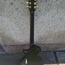Černá elektrická kytara s tvarem typu Les Paul, zobrazená z pohledu zadní strany. Na hlavě jsou zlaté ladičky a nápis "MADE IN KOREA". V horní části…