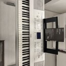Bílá digitální klaviatura s černými a bílými klávesami. Na přístroji je nápis YAMAHA a v pravém dolním rohu logo Clavinova. Vedle klaviatury leží čer…