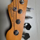 Detail hlavy basové kytary z přírodního dřeva. Na dřevě je černý nápis Fender, pod ním menší text MADE IN U.S.A. a vedle něj JAZZ BASS®. Nástroj má č…