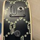 Detail zadní strany černého nástroje s krémovým lemováním. Povrch je zdoben motivem stylizovaných postav a čtverců v krémové barvě. Na nástroji je př…