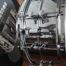 Detailní pohled na chromovaný malý buben s bílým koženem. Na mechanismu napnutí je viditelný nápis SONOR. Vedle nástroje je umístěna krabice s nápisy…