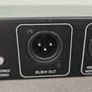 Detailní pohled na zadní panel černého elektronického zařízení. Jsou vidět dva konektory XLR, z nichž levý je označen nápisem SUBW OUT a pravý nápise…
