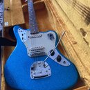 Modrá elektrická kytara s třpytivým povrchem v hnědém tvrdém kufru. Nástroj má stříbrný plechový kryt (pickguard) a na mostku je viditelný nápis Fend…