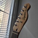 Fotografie zachycuje hlavu a horní část hmatníku elektrické kytary z přírodního dřeva s tmavým hmatníkem. Na hlavě je černý nápis Squier TELECASTER a…