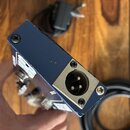 Detailní pohled na modrý kovový efekt pedál z úhlu. Na pravém boku je umístěn stříbrný konektor XLR s třemi piny. Zařízení je přišroubované čtyřmi če…