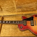 Elektrická kytara s červeně žlutým slupovaným povrchem korpusu v hnědém plišovém kufru. Na hlavě je nápis Epiphone a logo. Kytara má dva humbuckery,…