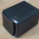 Černý, kostkovitý elektronický přístroj s matným povrchem. Na horní části je vyraženo logo ve tvaru stylizovaného obdélníku s vnitřními čarami. Na bo…