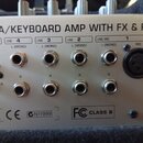Detail zadní strany stříbrného zesilovače s nápisem TT PA/KEYBOARD AMP WITH FX & FBQ. Jsou zde vidět vstupy CD IN (bílý a červený RCA), čtyři stereo…