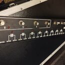 Detailní pohled na přední panel černého efektového přístroje v transportním kufru. Na horní části jsou čtyři kovové přepínače s popisky D, C, B a A.…