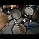 Elektronická bicí souprava s třemi bílými padami v černých rámech. Na padech jsou vidět nápisy Roland a V-Drums. K sadě patří kovový činel a dvě dřev…
