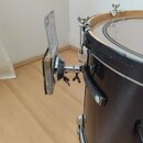 Detailní záběr na černý buben s připevněným kovovým držákem na činely. Držák je upevněn k obruchu bubnu pomocí šroubů a má stříbrnou barvu. Povrch bu…