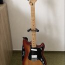 Elektrická kytara s tělem v červenohnědém sunburst provedení a světlým javorovým hmatníkem. Na hlavě je viditelné logo Fender a na popruhu nápis Fend…