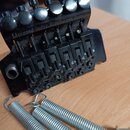 Černý kytarový kobylník s nápisem Licensed under Floyd Rose Parts. Součástí jsou tři stříbrné pružiny z kovového drátu. Povrch kobylníku vykazuje zná…
