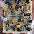 Fotografie znázorňuje elektronickou desku s osazenými součástkami, jako jsou rezistory, kondenzátory a tranzistory. Na desce je vidět černý nápis MXR…