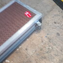 Fotografie zachycuje detail rohové části transportního kufru s hnědým, strukturovaným povrchem. Kufr má hliníkové hrany a v rohu je umístěno kolečko.…