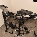 Elektronická bicí souprava v černé barvě s černými pady a činely. Na rámu je viditelný bílý nápis Roland. Souprava obsahuje ovládací modul s řadou tl…