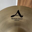Detailní pohled na mosazný činel. Na povrchu je černý nápis s logem v podobě stylizovaného písmene A a textem MEDIUM CRASH 18"/45 cm. Povrch činelu v…