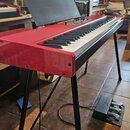 Červené digitální piano na černém kovovém stojanu s přívodním kabelem. Součástí je černý notový pult a černý pedál s třemi st expressesními pedály, n…