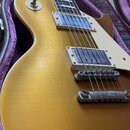Detail elektrické kytary typu Les Paul v zlatavé barvě, která leží v otevřeném kufru s fialovou výstelkou. Na těle jsou dva snímače v bílých rámečcíc…