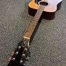 Akustická kytara s přirozeným dřevěným povrchem korpusu a černou hlavou. Na hlavě je zlatý nápis Epiphone a logo v horní části hmatníku. Nástroj vyka…