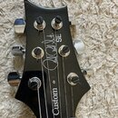 Detail hlavy černé elektrické kytary s chromovanými ladičkami. Na povrchu je bílý nápis „Paul Reed Smith SE“ a svisle orientovaný nápis „Custom“. Lak…