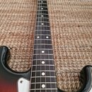Elektrická kytara s tmavým hmatníkem a bílými tečkovými značkami poloh. Tělo má černo-oranžový přechodový lak a bílý pickguard. Na hlavě krku je vidi…