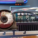 Detailní pohled do otevřeného elektronického zařízení. Je vidět velký toroidní transformátor s červeným jádrem a bílým papírovým štítkem s nápisem PA…