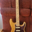 Elektrická kytara typu Stratocaster s přírodním dřevěným povrchem těla a hmatníkem. Tělo má tmavě červený želvový pickguard a tři bílé snímače. Na hl…