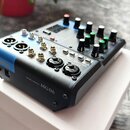 Černý mixážní pult z kovového a plastového materiálu s modrými detaily. Na předním panelu jsou nápisy MIXING CONSOLE MG06 a YAMAHA. Zařízení obsahuje…