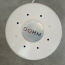Bílé kulaté zařízení z plastu s otvory v horní části. Uprostřed je stříbrný kruh s nápisem DOHM. Kolem něj je vypsáno ORIGINAL NATURAL WHITE NOISE MA…