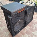 Dvě černé basové reprobedny s kovovými mřížkami, na kterých je umístěn rackový zesilovač. Zesilovač má oranžový nápis the t-amp TA 1400 a pod ním men…