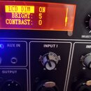 Detailní záběr na ovládací panel elektronického zařízení s červeným displejem, na kterém je text LCD DIM: ON, BRIGHT: 5 a CONTRAST: 0. Pod displejem…