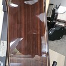 Detail zadní strany akustické kytary s hnědým lakovaným povrchem z dřeva. Nástroj je umístěn v černém stojanu, na kterém je viditelný nápis "stand" a…