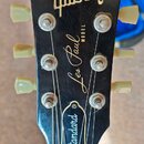 Černá hlava elektrické kytary se značkou Gibson. Na hlavě jsou nápisy Les Paul MODEL a Standard. Bílé ladičky vykazují známky opotřebení a povrch hla…