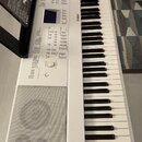 Bílá digitální klaviatura značky YAMAHA s integrovanými reproduktory a ovládacím panelem. Na levé straně je umístěn černý stojan na noty s vloženým p…