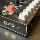 Černý elektronický instrument s bílými a černými klávesami a jednou červenou klávesou. Na boku jsou tři konektory a nápis "chomp club". Povrch je čer…