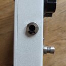 Detailní pohled na boční stranu bílého efekt pedálu s kovovým povrchem. Na boku je umístěn jeden kovový vstupní/výstupní konektor typu jack. V horní…