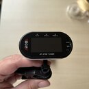 Černý digitální ladicí přístroj s připevněným klipem. Na přední straně je displej a nápisy FLAT, PITCH, ITEM/電源 s ikonami šipek, vlevo červené logo A…