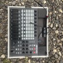Černý MIDI kontrolér Novation APC40 MKII umístěný v otevřeném hliníkovém kufru na štěrku. Zařízení obsahuje mřížku podsvícených padů, posuvníky a něk…