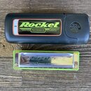 Fotografie zobrazuje foukací harmoniku v zeleném plastovém rámu s kovovými kryty, položenou vedle černého plastového pouzdra. Na harmonice i na nálep…