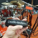 Černý plastový držák nebo kryt s bílým nápisem audio-technica® a doprovodným logem. Předmět drží ruka osoby s černými hodinkami na zápěstí. V pozadí…