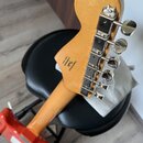 Záběr na zadní stranu hlavy a krku elektrické kytary v přirozené dřevěné barvě. Na zadní straně hlavy je černý nápis Designed by Fender s logem Fende…