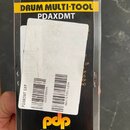 Fotografie znázorňuje produkt v originálním černém papírovém obalu. Na horní části je žlutý pruh s nápisem DRUM MULTI-TOOL a pod ním model PDAXDMT. V…