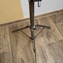 Kovový stativ s trojnožkou umístěný na dřevěné podlaze. Povrch kovových trubek vykazuje výrazné známky opotřebení, korozi a odřeniny. Na stativu není…