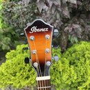 Fotografie zachycuje hlavu akustické kytary s barevným přechodem od oranžové k černé. Na horní části je bílý nápis Ibanez. Kytara má šest černých lad…