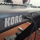 Černý klávesový nástroj s bílými a černými klávesami. Na zadní straně je velký bílý nápis KORG a vedle něj menší nápisy KRONOS a MUSIC WORKSTATION. D…