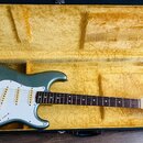 Elektrokytara s modrozeleným trupem a bílým pickguardem v černém kufru s žlutým polstrováním. Na hlavě hmatníku je vidět logo Fender a nápis STRATOCA…